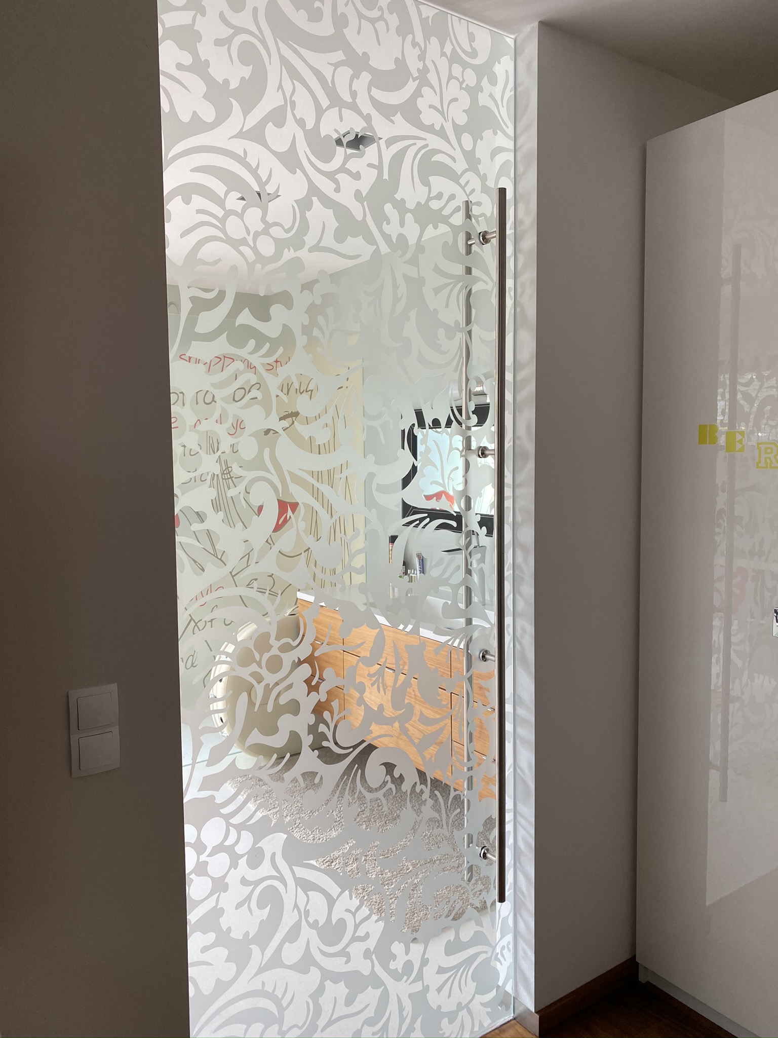 Satinierte Glastür mit floralem Ornament und langem Metallgriff; dahinter ein heller Raum mit Holzkommode und Spiegel.