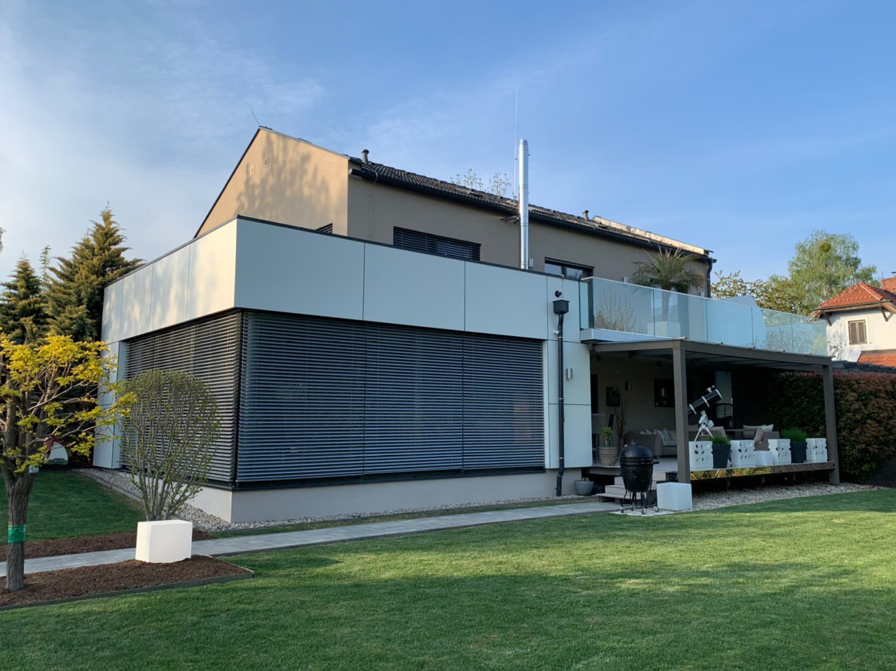 Modernes Wohnhaus mit weißem Flachdach-Anbau, geschlossenen Außenjalousien, Glasbalkon und überdachter Terrasse mit Sitzbereich; davor gepflegter Rasen und Garten unter blauem Himmel.
